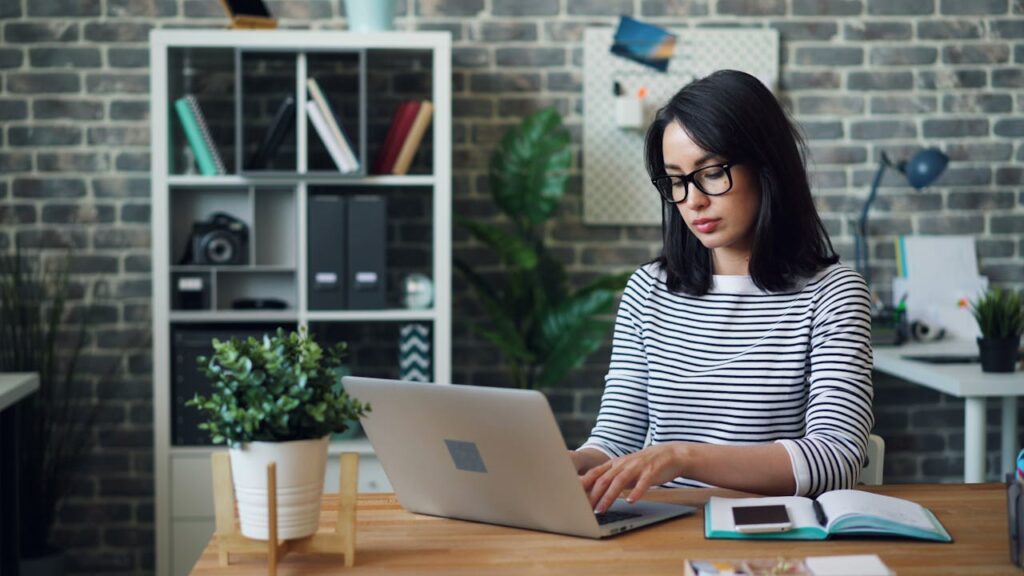 Professional woman working on her laptop in a modern office setting, surrounded by books and plants.