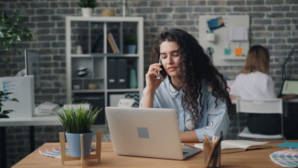 Young woman multitasking at her desk with a laptop and phone in a stylish office.