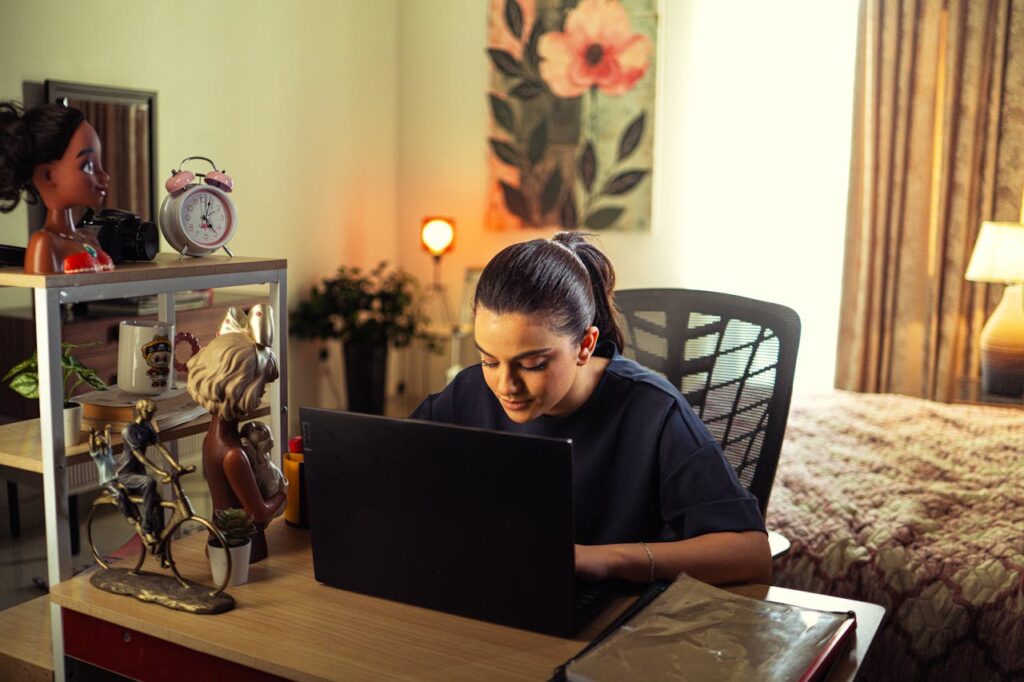 A woman focusing on her laptop in a cozy home office surrounded by decor and warm lighting.